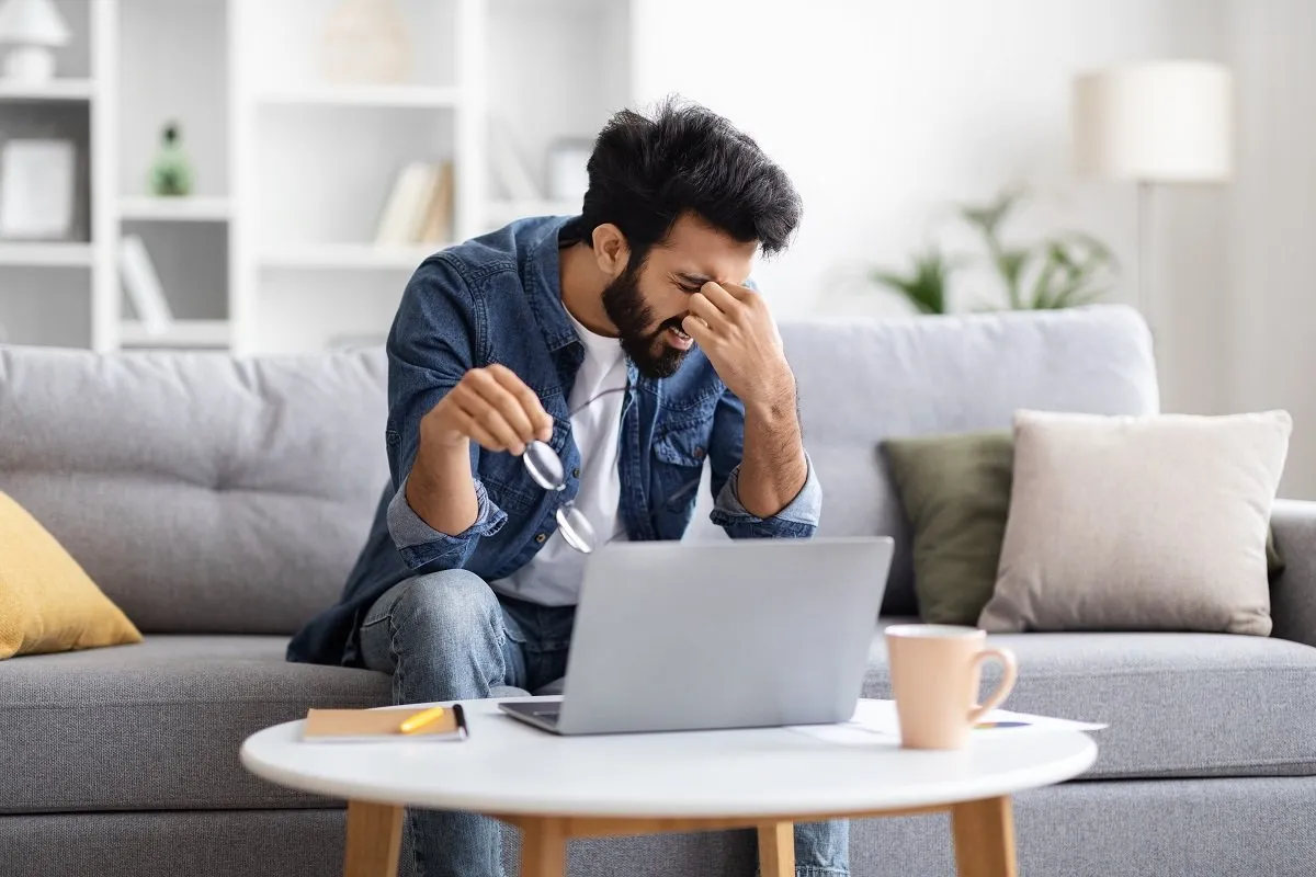 A frustrated man holding glasses near the laptop.