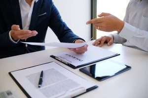 Two people with their heads out of frame are around a table with clipboards and a tablet on it. The one on the left is holding a piece of paper while the one on the right gestures to it.