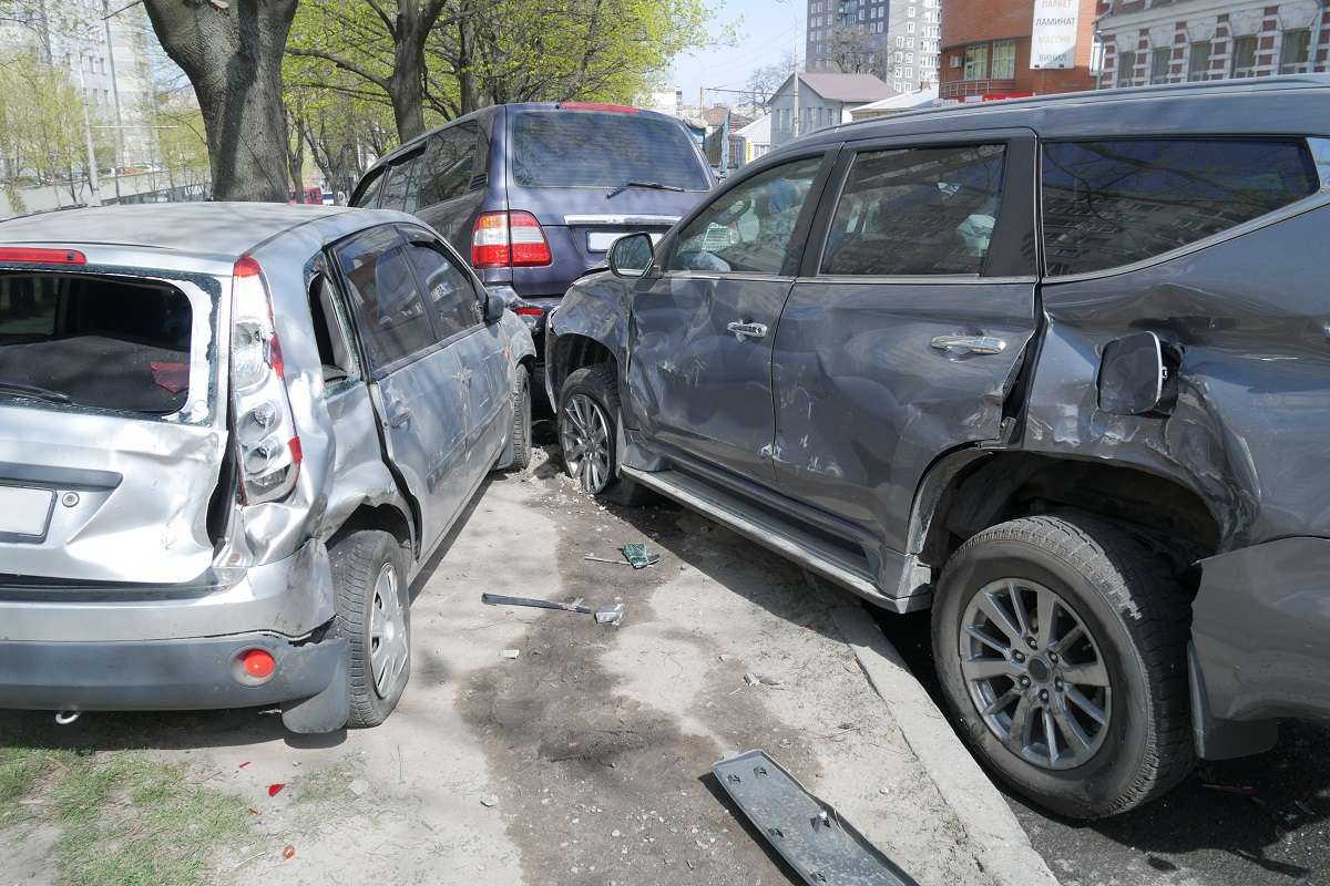 A small silver car and a dark SUV are both crashed in the back of a blue vehicle.