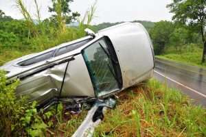A damaged silver passenger van is on its side in an overgrown area.