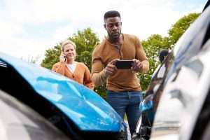 A person wearing blue takes a photo of the damage of two cars while another person makes a call.