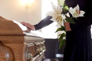 A person wearing black and holding lilies has a hand on a casket.