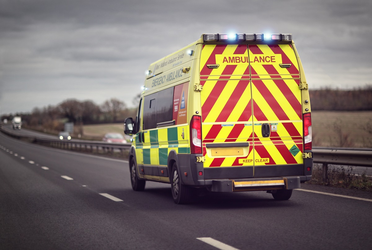 An ambulance with yellow and ride stripes on the back is driving down the road with its lights on.