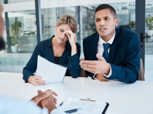 A person sits across from a person growing and holding a sheet of paper next to another in a suit and talking.