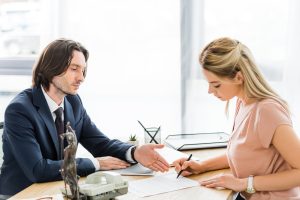 A person in a suit gestures at a document another person in a pink shirt is signing.
