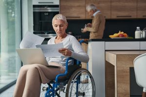 A person in a wheelchair sits in front of a kitchen and is frowning at pieces of paper.