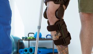A person with a knee brace and crutches is shown from the knee down. A second person in blue scrubs is on the left.