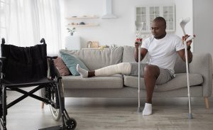 A man sits on a couch with a bandaged leg. He is holding crutches on the ground and there is a wheelchair nearby.
