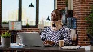 A man wearing a cervical collar is seated at a desk and looking at a laptop.