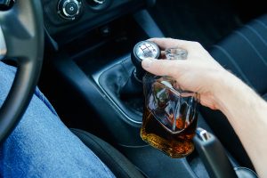 A close-up of a driver reaching for a stick shift while holding an open bottle of alcohol.