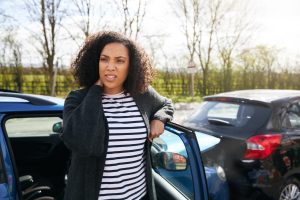 A woman stands leaning against her open car door, holding her neck in pain after a rear-end car accident.