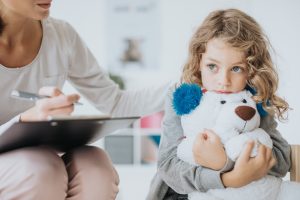 A sad little girl with a teddy bear sits next to an adult with a clipboard.