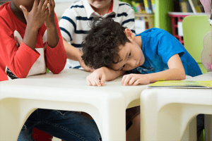 A sad little boy sitting at a school table with his head resting on his arm